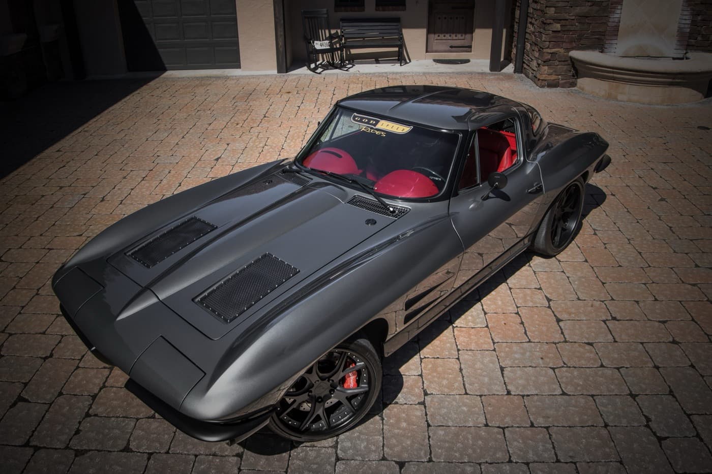 A matte grey split-window Corvette with a red interior and black wheels, shot from above on a brick driveway