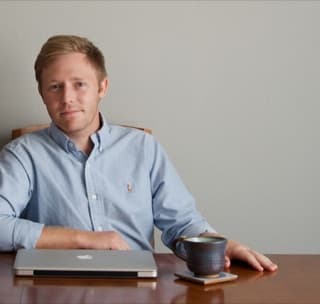 Brandon Selfors in a light blue shirt at a wooden table, a laptop and a cup of coffee in front of him