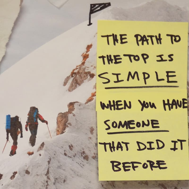 A yellow sticky note hand-lettered “The path to the top is simple when you have someone that did it before”, stuck over a photograph of two roped climbers walking a snow ridge