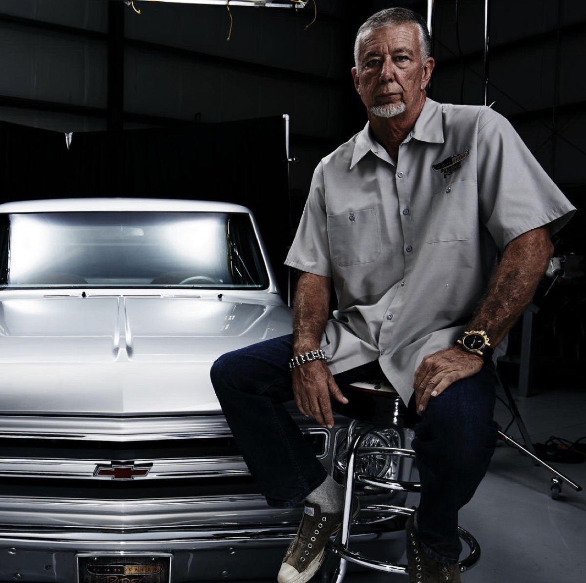 A grey-haired man in a work shirt sitting on a stool beside the polished chrome front end of a classic Chevrolet pickup in a dark workshop
